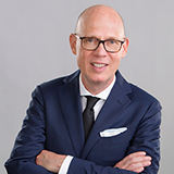 Carsten Jensen, a man with close-cropped hair and glasses, poses with hims arms crossed wearing a blue suit