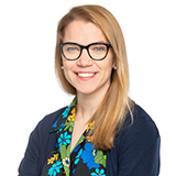 Anna Lund, a woman with long blonde hair and glasses, poses wearing a blue cardigan over a colourful blouse.