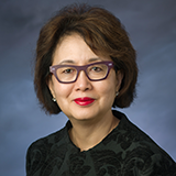 Lillian Pan, a woman with short dark hair and glasses, poses wearing a dark blazer in front of a blue background.