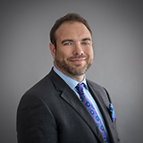 David Benjestorf, a man with brown hair and a beard, poses in front of a gray background wearing a dark suit and blue tie.