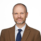 Eric Adams, a man with close cropped hair and a gray beard, poses wearing a brown suit jacket and blue tie