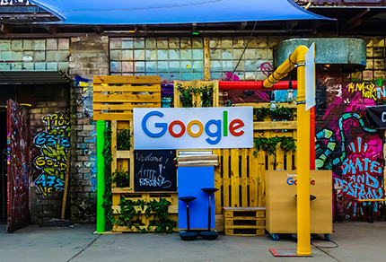 A decorated area with a large Google sign, wooden pallets, green plants, and graffiti-covered walls in the background.