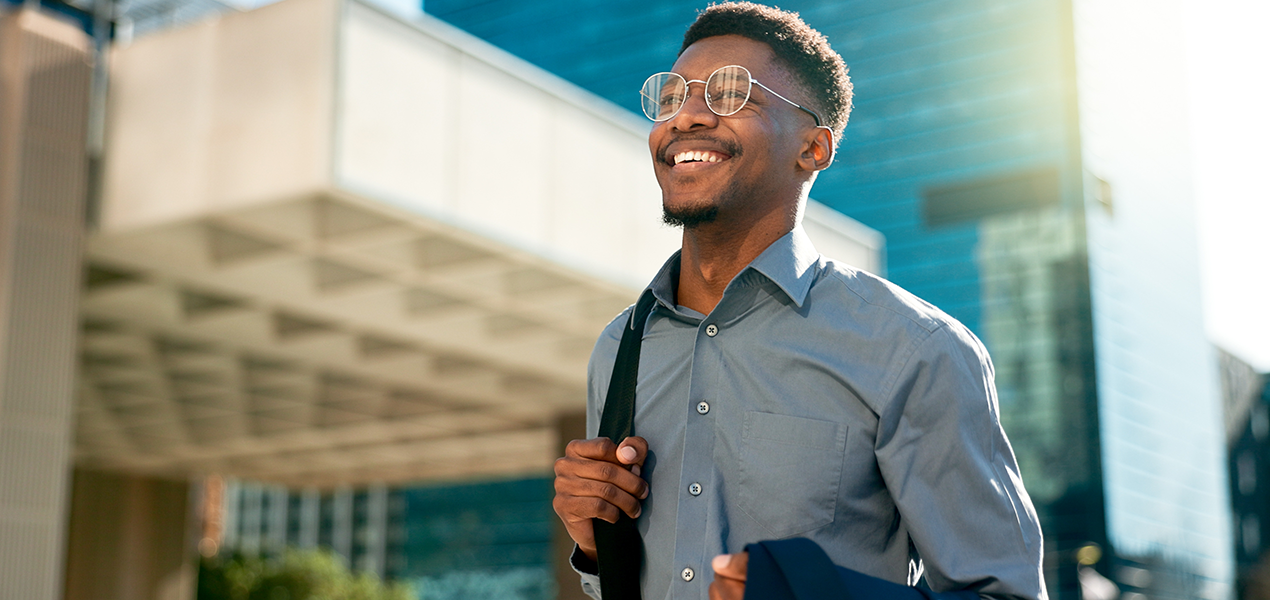 A professional Black man wearing glasses and a button-down shirt smiles while walking outdoors in an urban area on a sunny day.