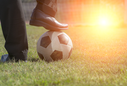 close-up of a person’s foot resting on a soccer ball on a grassy field, with sunlight in the background.