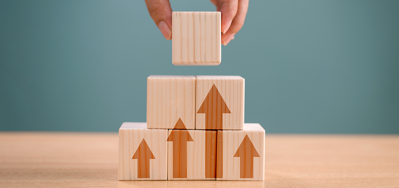 A hand places a wooden block on top of a stack of blocks with upward-pointing arrows, symbolizing growth or progress.
