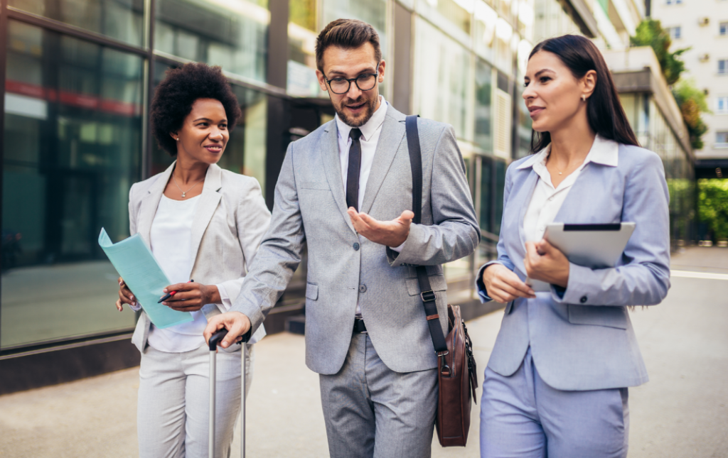 Three business professionals walk outside an office building, conversing. One holds a tablet, one carries a briefcase and gestures, and one pulls a suitcase while holding documents.