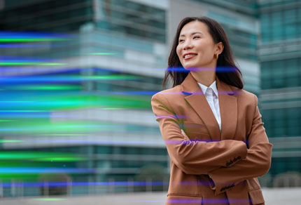 Confident woman in a tan blazer standing with arms crossed in front of modern glass office buildings, with blurred green and blue light trails passing across the foreground.