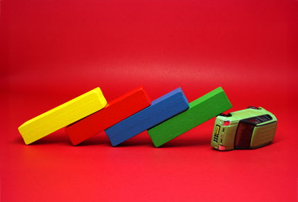 A toy bus is tipped over with four colored blocks—yellow, red, blue, and green—stacked and falling like dominoes against it, set on a red background.