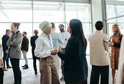 A group of people wearing business attire stand in a modern office space, talking and networking in small groups.