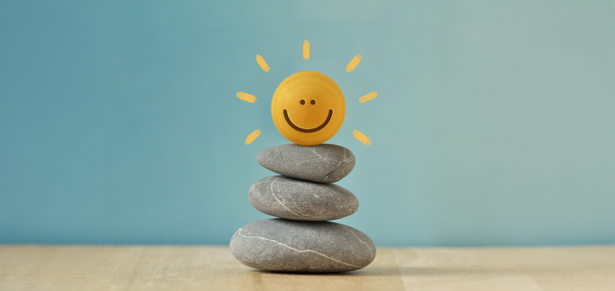A minimalist scene of three smooth gray stones stacked on a wooden surface, topped with a small yellow ball painted with a smiling face. Simple sun rays are drawn around the ball, set against a soft b