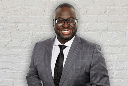 Mosi Taylor-Cole, a Black man with short hair and glasses, poses in front of a white brick wall wearing a grey suit.