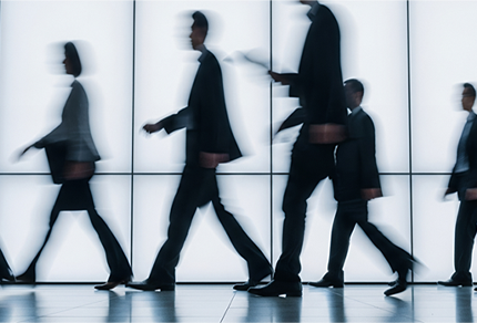 Blurred silhouettes of business professionals walking past a glass wall, suggesting movement and anonymity