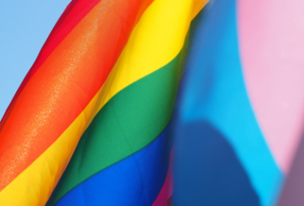 Close-up of a rainbow Pride flag and a pink, blue, and white transgender Pride flag against a clear blue sky.