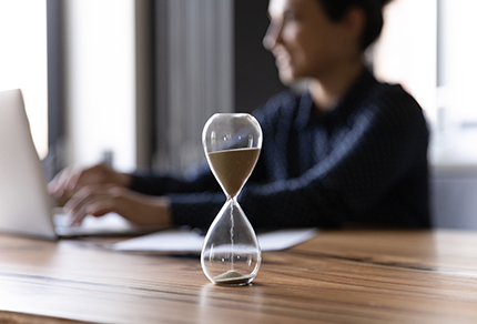 A professional woman in a suit working on a laptop, with an hourglass in the foreground.