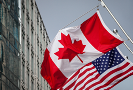 The Canadian and American flags are flying side by side on flagpoles outside a modern building.