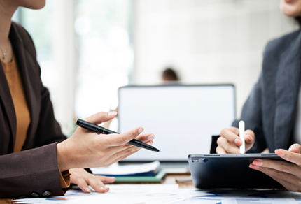 Two people in business attire discussing documents; one holds a pen and paper, the other uses a stylus on a tablet. A laptop is open on the desk in the background.